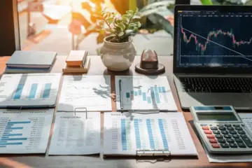 Financial documents, calculator, and laptop with stock chart on a wooden desk with a small plant.