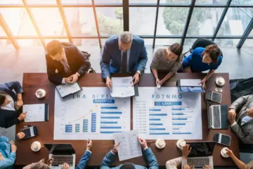 Diverse group at office table reviewing documents and charts with laptops and calculators.