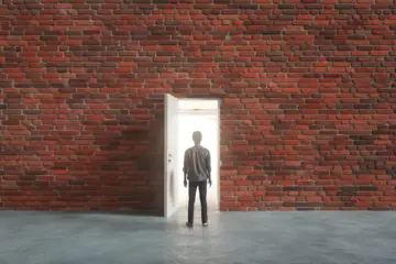 Person facing a brick wall with a small door ajar, showing a bright room behind it.