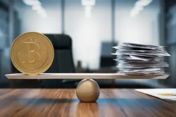 Scale with a gold coin on one side and paperwork on the other, on a desk with a blurred office background.