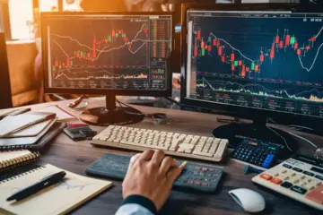 Desk with monitors showing stock charts and financial data, surrounded by pens, notebook, and calculator.