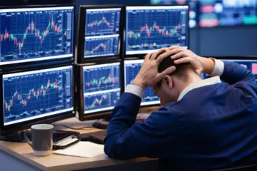 Trader at desk with multiple monitors showing stock charts, coffee mug nearby, hands on head in stress.